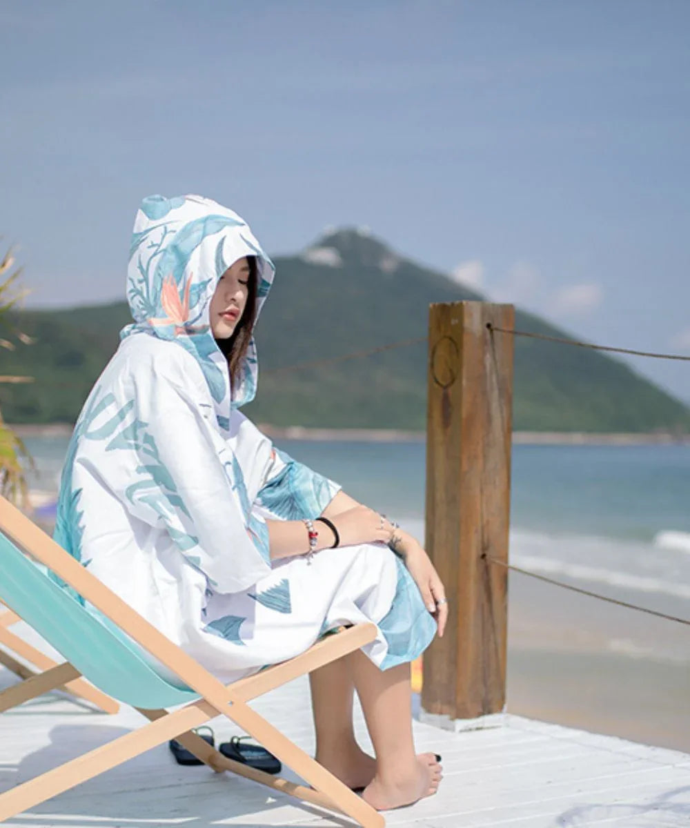 Femme en poncho surf blanc à motifs tropicaux sur une plage, assise près de la mer