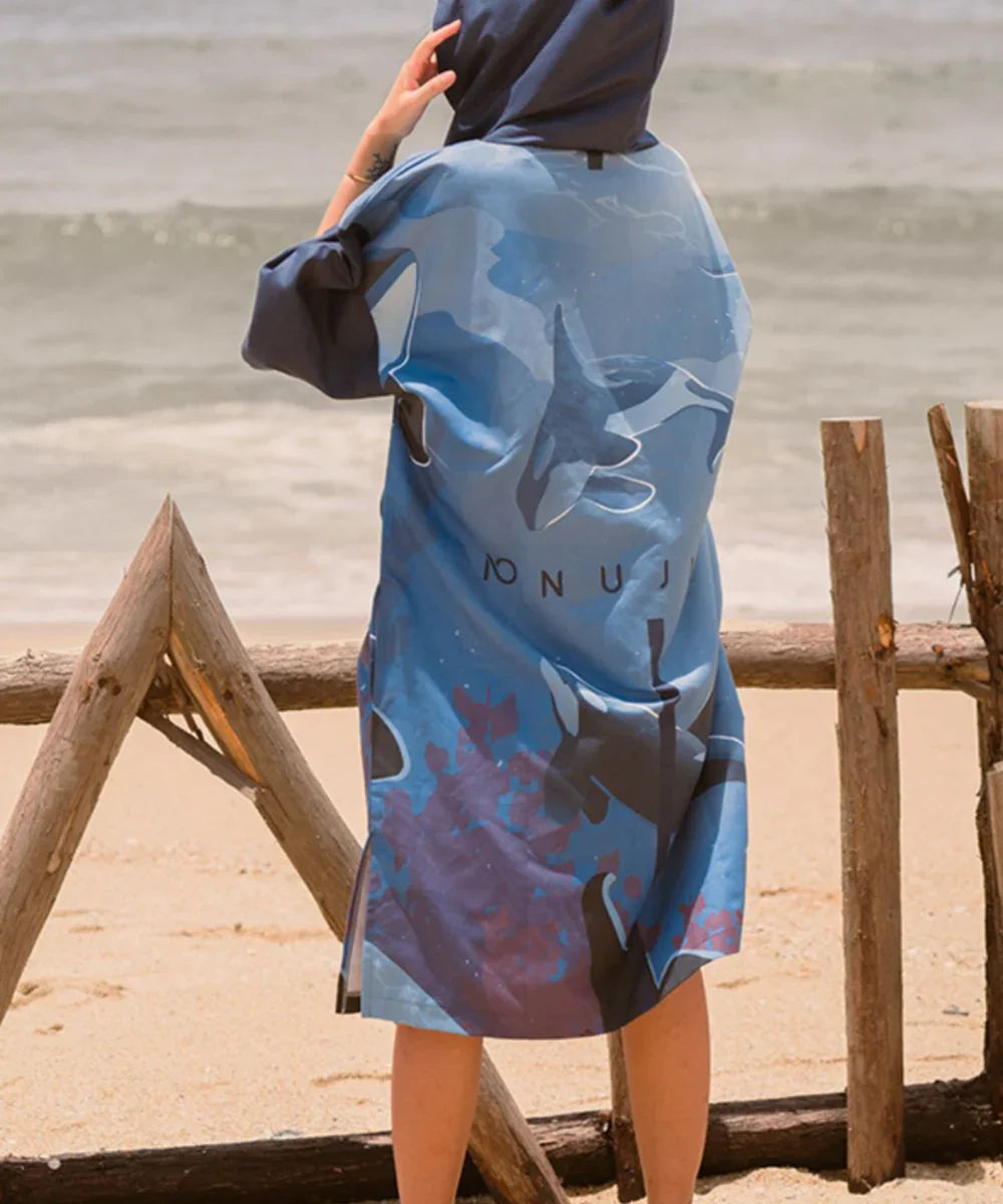 Personne portant un poncho surf à manches longues bleu avec capuche, motif orque, sur la plage