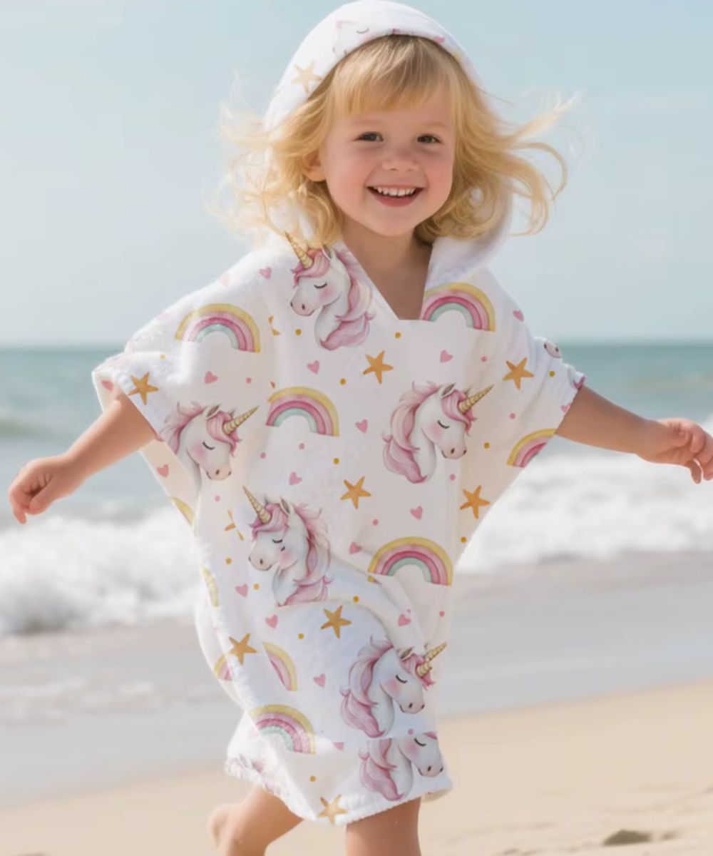 Petite fille souriante portant un poncho serviette à capuche Planète Poncho avec motifs licornes.